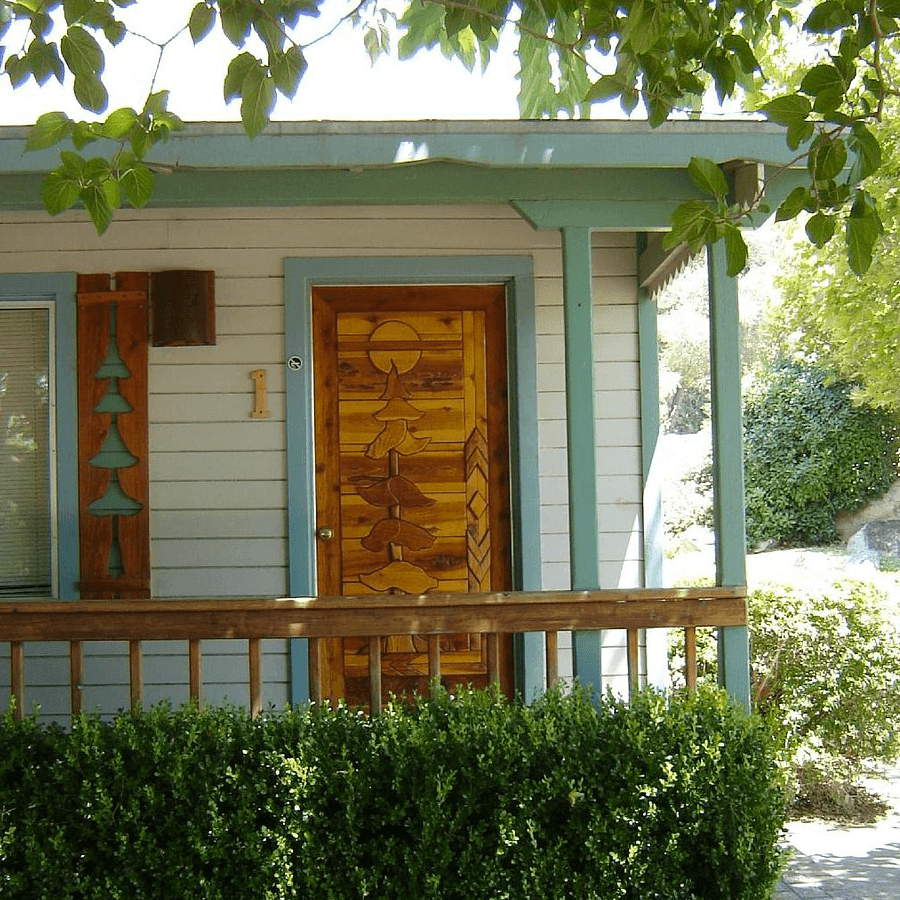 A colorful, intricately designed wooden door is framed by a light blue house with green foliage nearby.