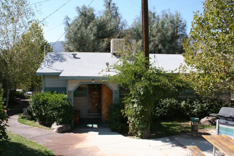 A small, blue-green house surrounded by greenery and trees.