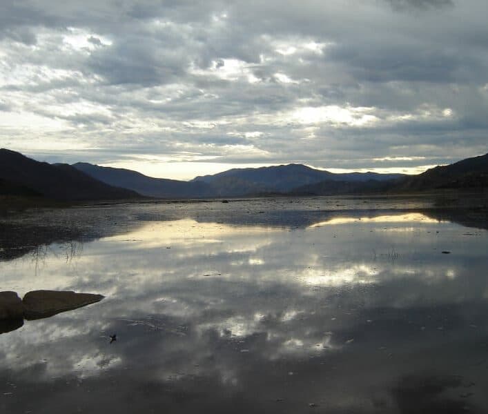 A calm lake reflects the cloudy sky and surrounding mountains.