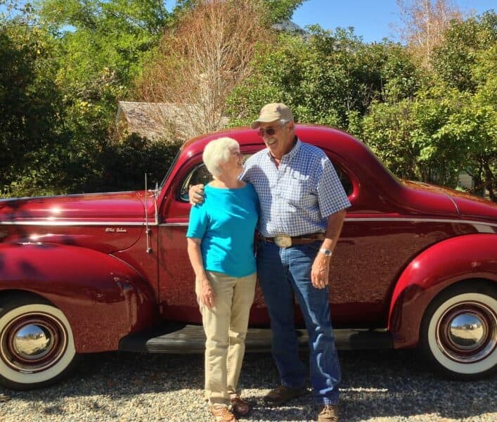 An elderly couple smiles at each other in front of a vintage red car.